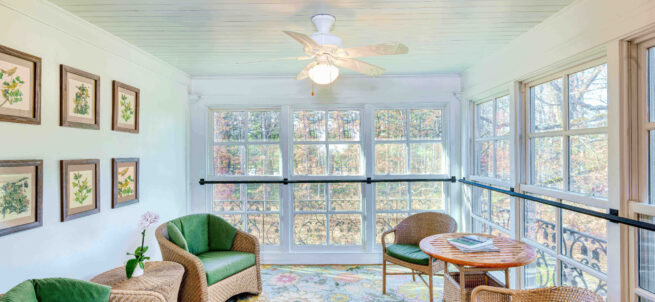 Bright sunroom with wicker furniture, green cushions, floral rug, and large windows overlooking trees.