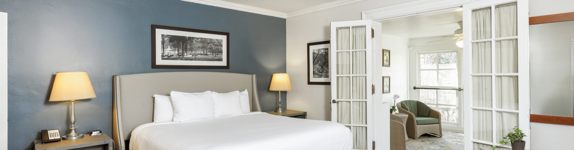 Modern hotel room with a large bed, two lamps, and open French doors leading to a bright sitting area.