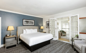 Modern hotel room with a large bed, two lamps, and open French doors leading to a bright sitting area.