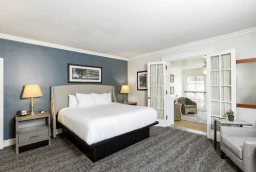Modern hotel room with a large bed, two lamps, and open French doors leading to a bright sitting area.