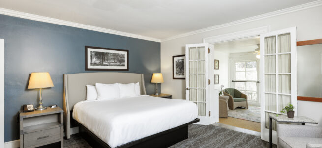 Modern hotel room with a large bed, two lamps, and open French doors leading to a bright sitting area.