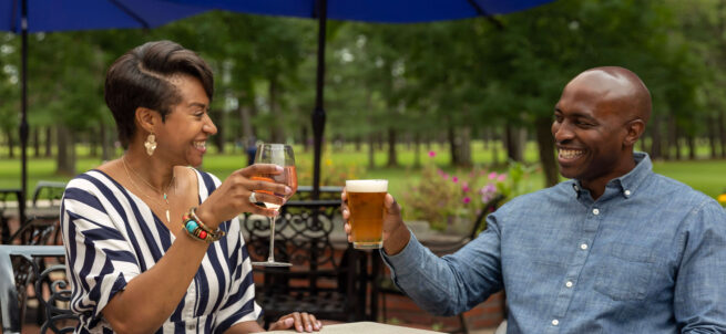 A woman and man sit outdoors, smiling and clinking a wine glass and beer glass together.