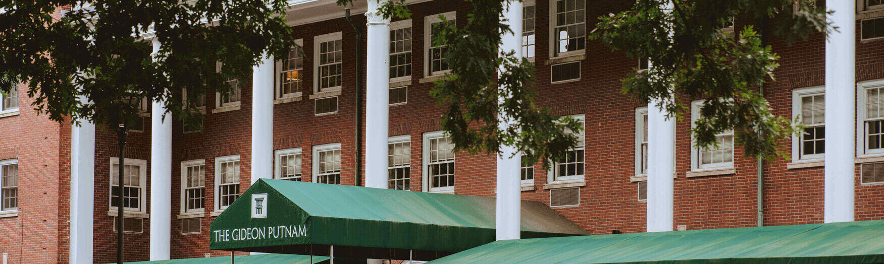 Historic red-brick hotel with white columns and green awnings, surrounded by potted plants and outdoor seating.