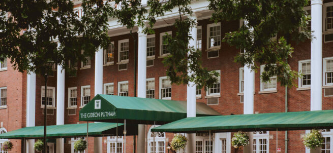 Historic red-brick hotel with white columns and green awnings, surrounded by potted plants and outdoor seating.