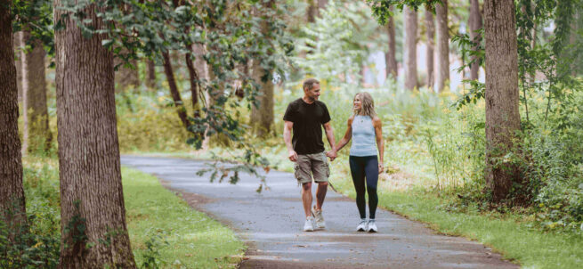 A couple holds hands while walking on a tree-lined path in a green park.