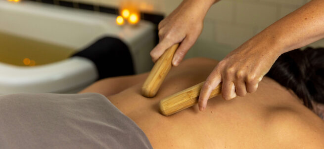 Person receiving a bamboo massage on their back in a spa setting with lit candles in the background.