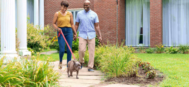 A couple walks a small dog on a leash along a garden path in front of a brick building.