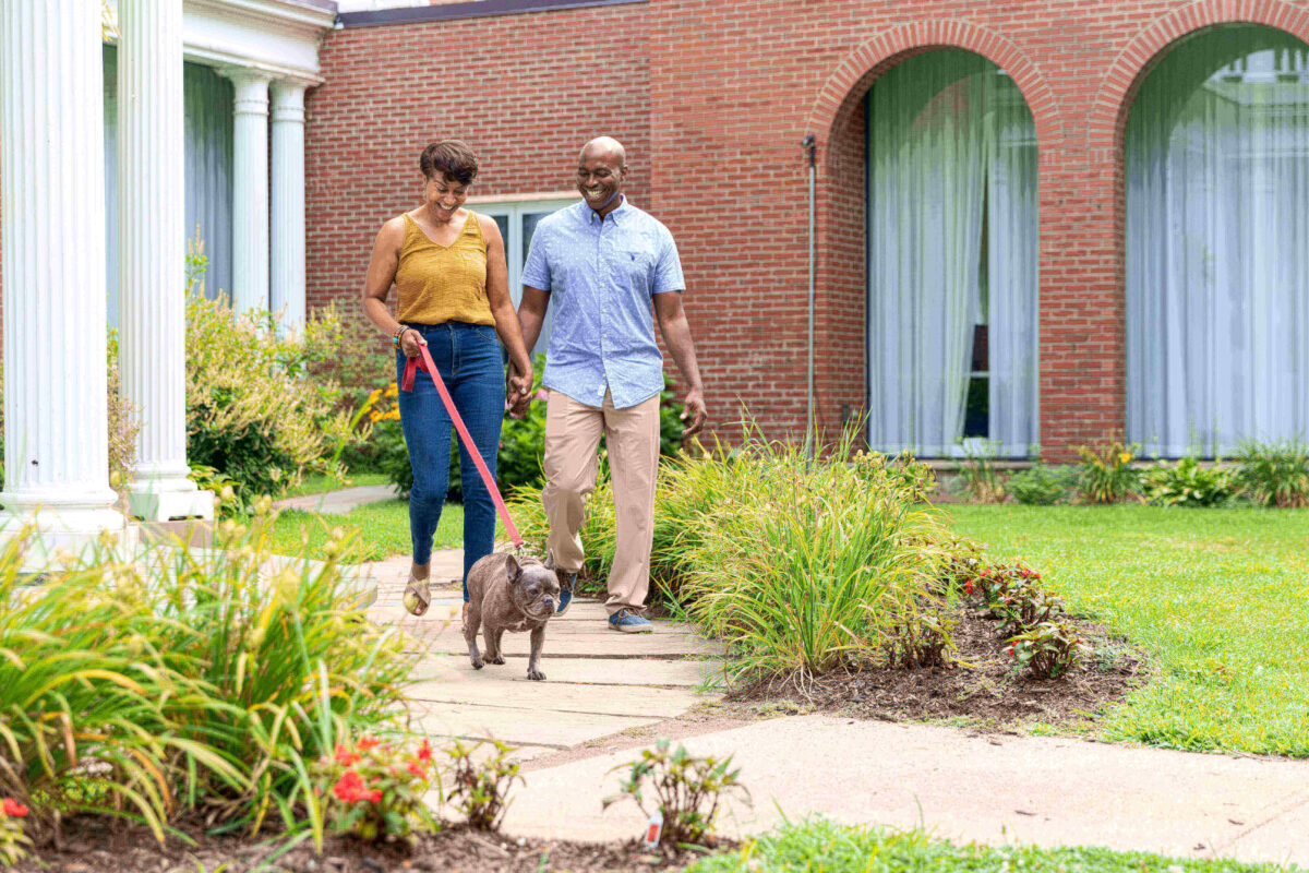 A couple walks their dog on a leash along a garden path in front of a brick building.