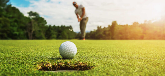 A golf ball near the hole with a golfer putting in the blurred background on a sunny day.