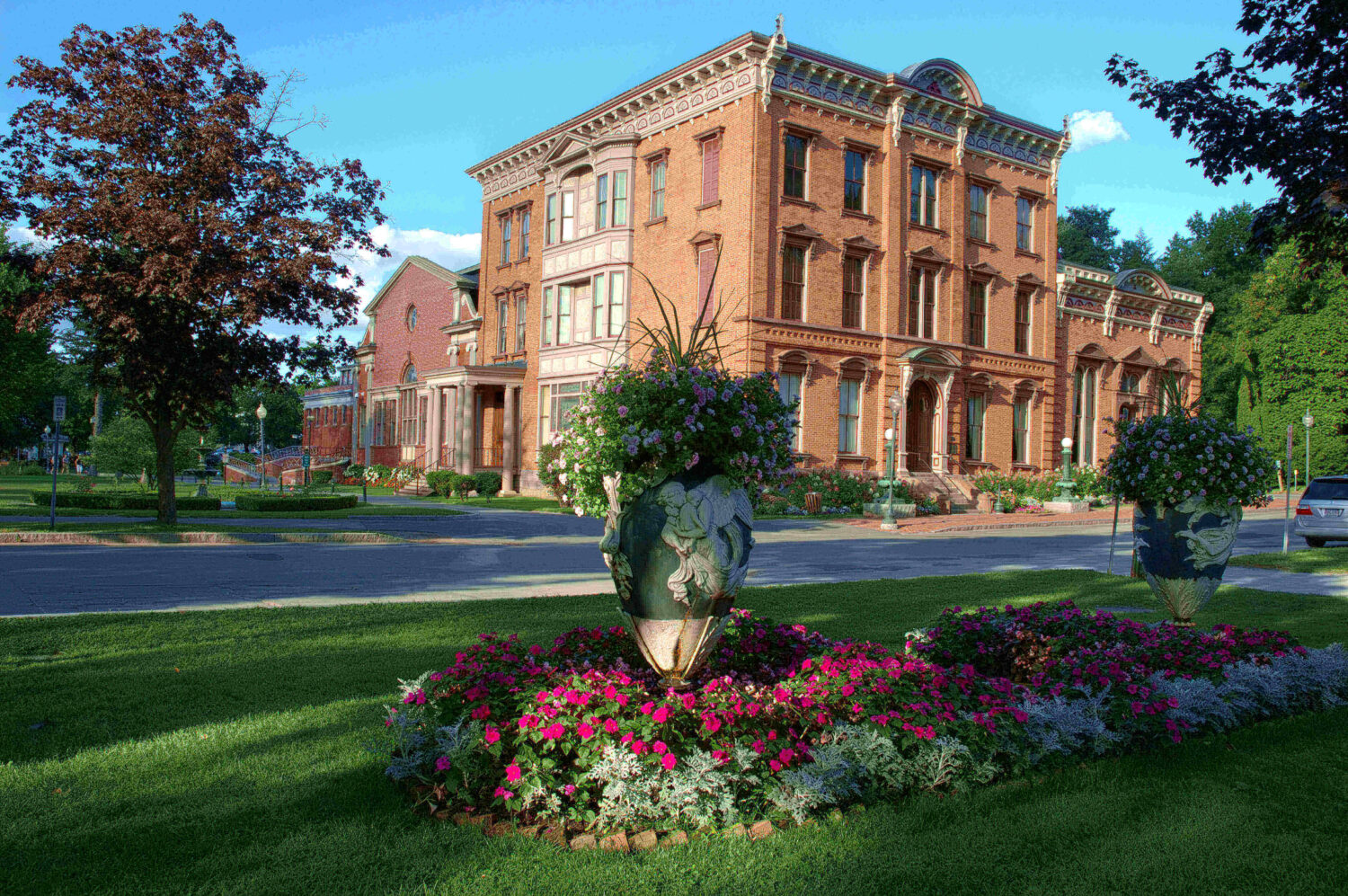 Red brick historic building with large flowerpots and colorful blooms on a sunny, green lawn.