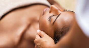 A woman lying down with eyes closed, receiving a relaxing head massage.