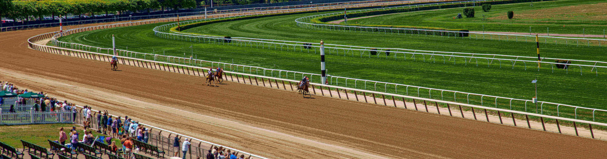 Horse race in progress on a dirt track, with spectators watching from the stands under a sunny sky.