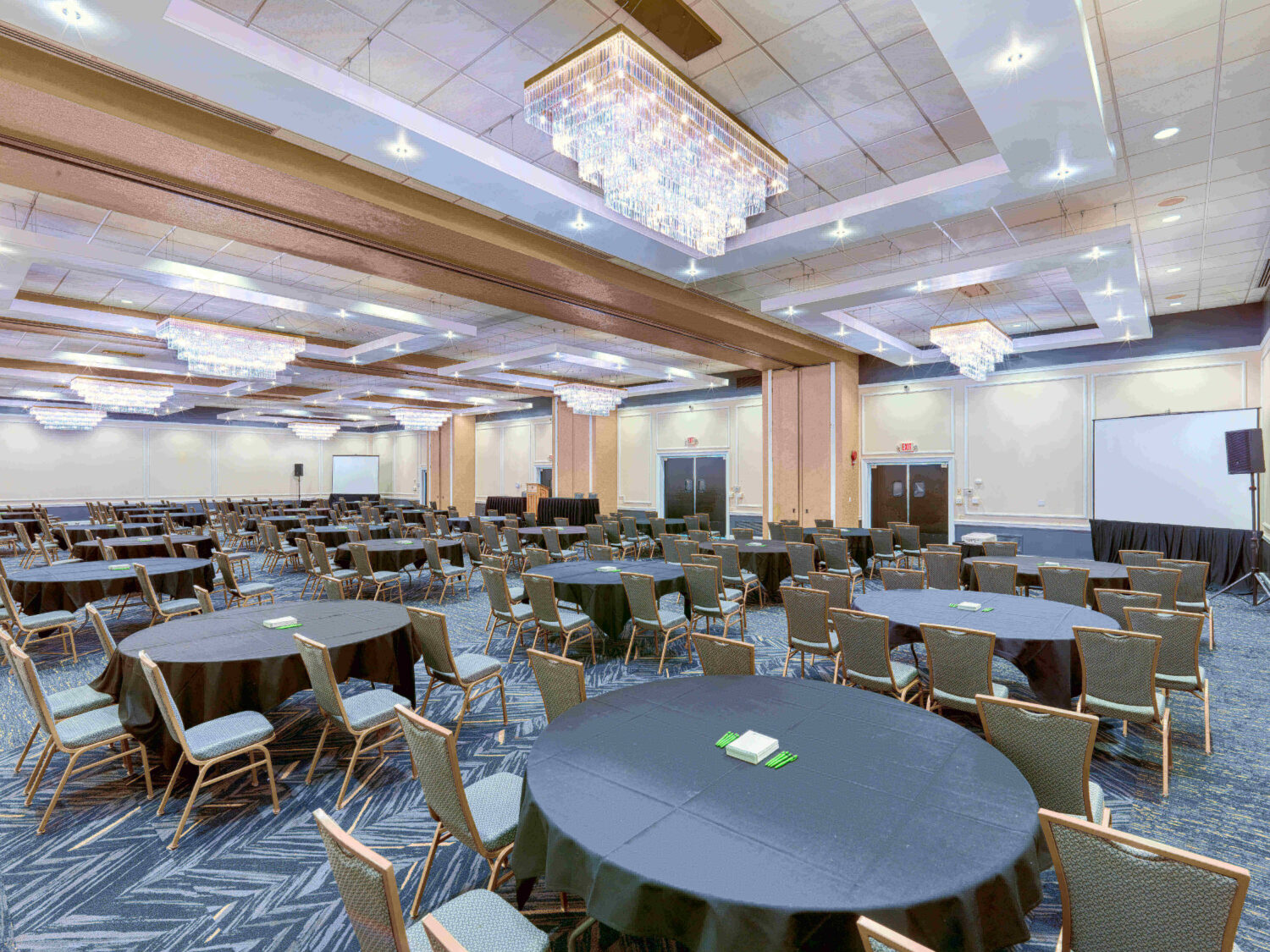 Large, empty conference room with round tables, chairs, chandeliers, and presentation screens.