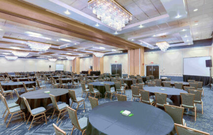 Large, empty conference room with round tables, chairs, chandeliers, and presentation screens.