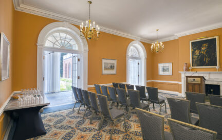 Rows of chairs face a fireplace in an elegant room with orange walls, chandeliers, and arched doorways.