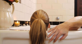 Person with blonde hair in a ponytail relaxing in a bathtub, with candles and flowers nearby.
