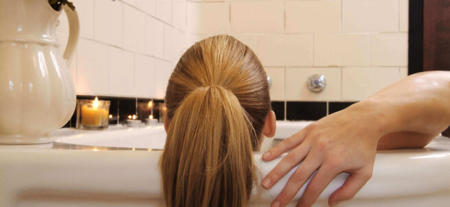 Person with blonde hair in a ponytail relaxing in a bathtub, with candles and flowers nearby.