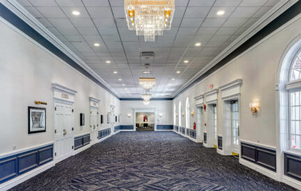 Spacious, elegant hallway with chandeliers, blue patterned carpet, white walls, and framed artwork.