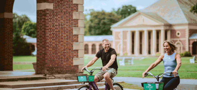 Two adults ride rental bikes on a path near a brick building and a large columned structure.