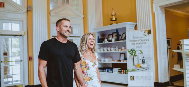 Smiling couple holding hands and walking inside a bright, elegant spa lobby with yellow walls.