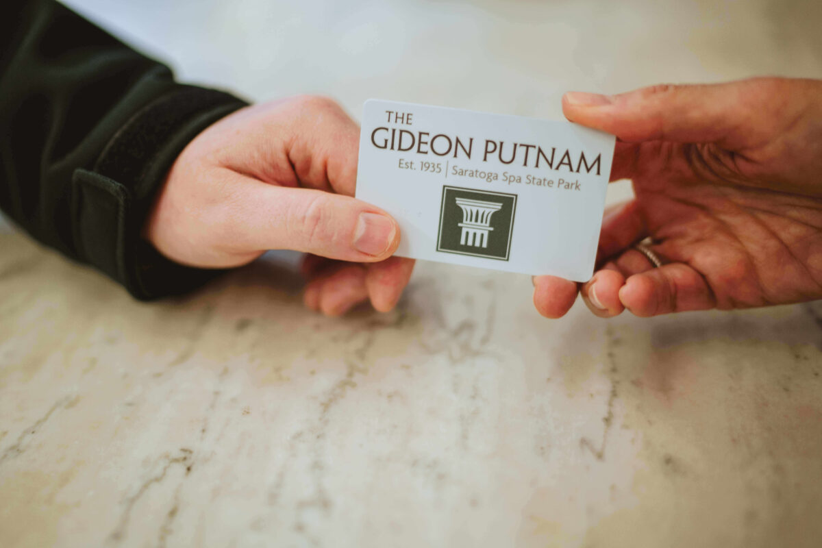 Two hands exchange a card labeled The Gideon Putnam, Saratoga Spa State Park over a marble surface.