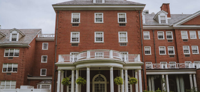Large red-brick building with white columns, balconies, and green plants, seen from a grassy lawn.