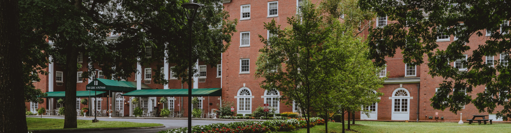 Red-brick building with white windows, surrounded by trees, flowers, and curved sidewalks.