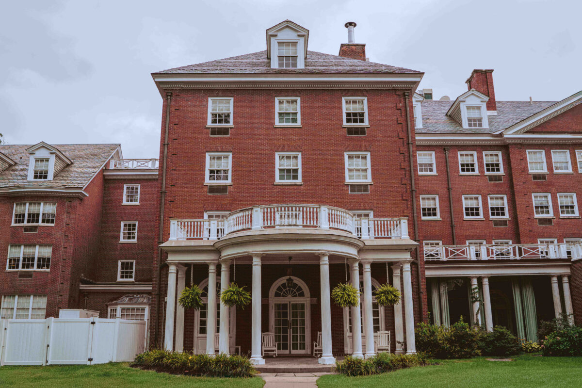 Large red-brick building with white columns, balconies, and many windows, surrounded by green grass.