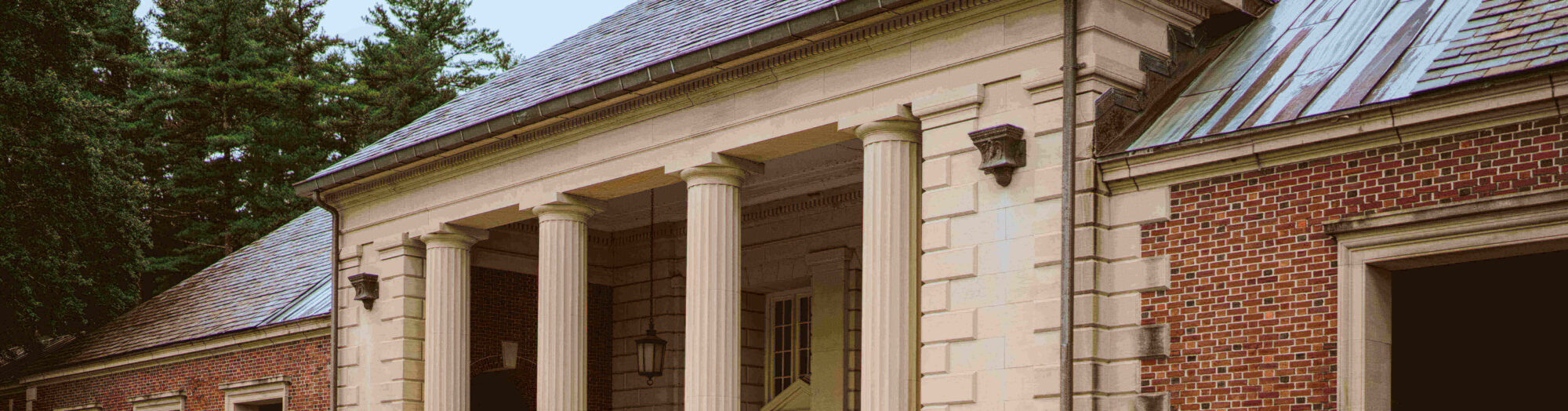 A historic brick building with four tall columns at the entrance and a peaked roof, surrounded by trees.
