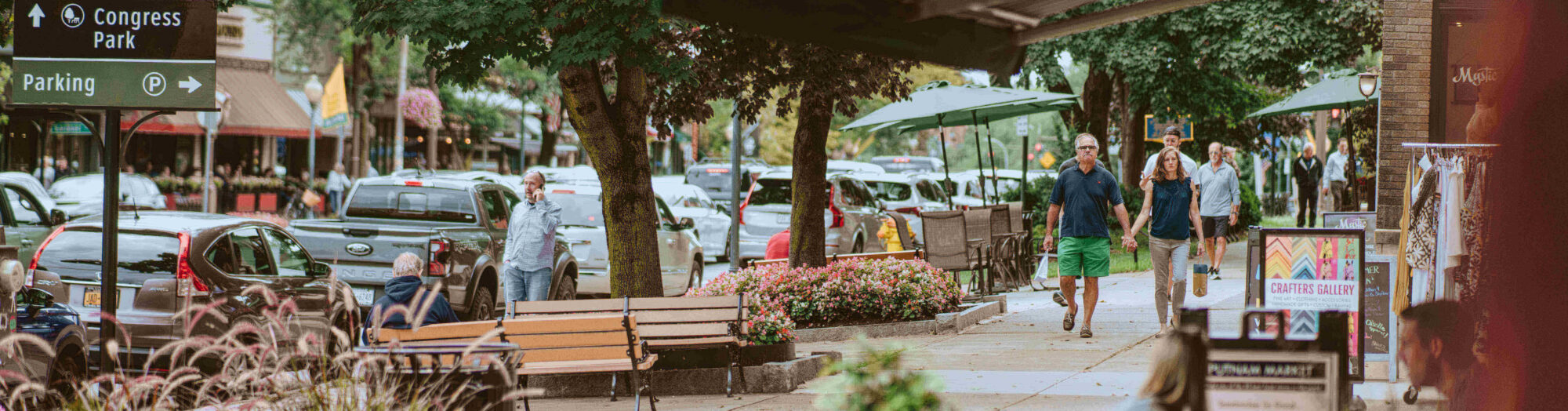 People walk along a lively sidewalk with shops, benches, and parked cars on a tree-lined street.