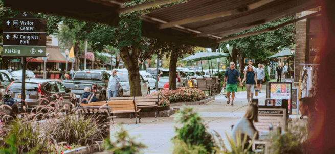 People walk along a lively sidewalk with shops, benches, and parked cars on a tree-lined street.