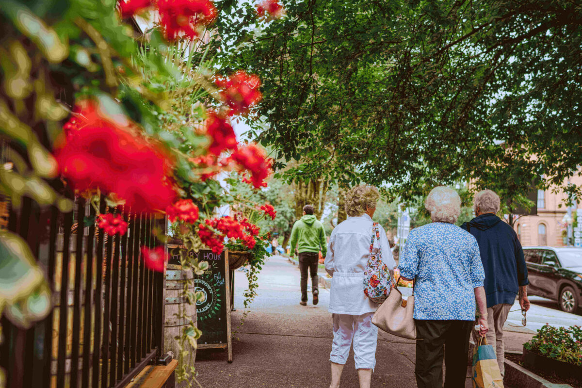 Four older adults walk on a tree-lined sidewalk with red flowers in the foreground.