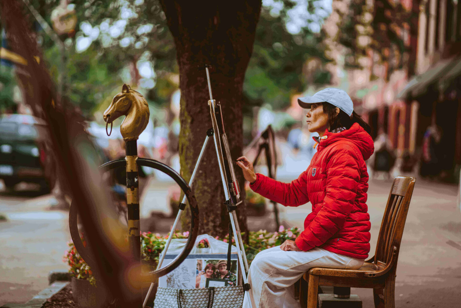 Woman in a red jacket painting outdoors, seated by an easel on a city sidewalk near a horse-shaped post.