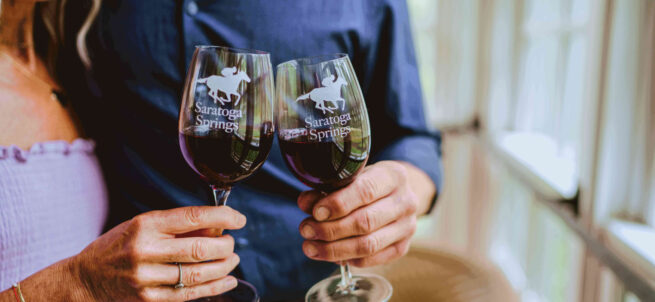 A couple clinks Saratoga Springs wine glasses together, each holding a glass of red wine.
