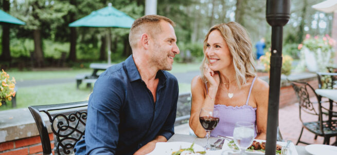 A smiling couple enjoys a meal and wine together at an outdoor restaurant patio.