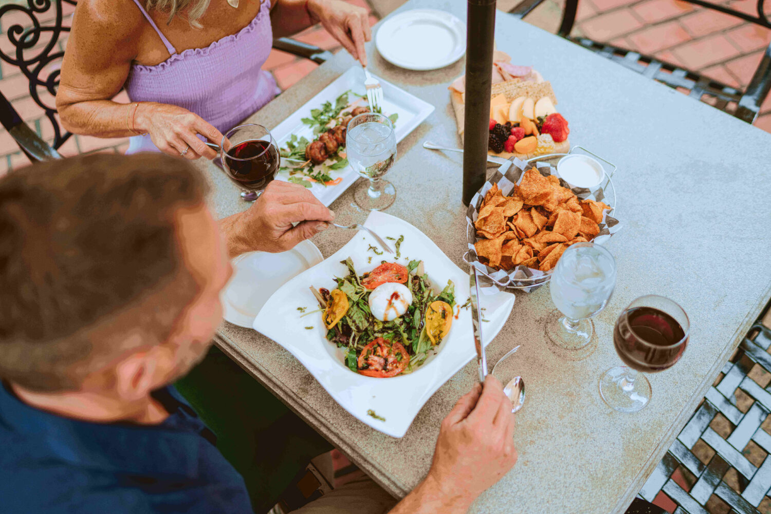 Two people dining outdoors with wine, salads, fruit, and chips on a stone table.