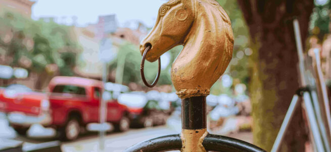 A gold horse-head hitching post on a sidewalk, with cars and trees blurred in the background.