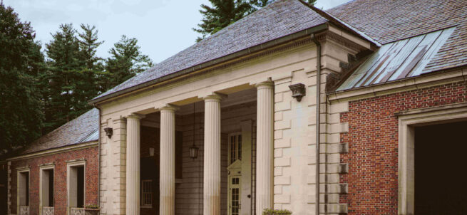 A brick mansion with tall white columns at the entrance, surrounded by trees and cloudy sky.