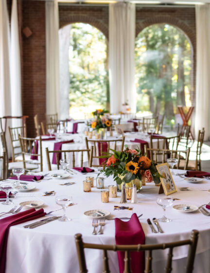 Round tables with white tablecloths, burgundy napkins, and floral centerpieces in a bright, elegant room.