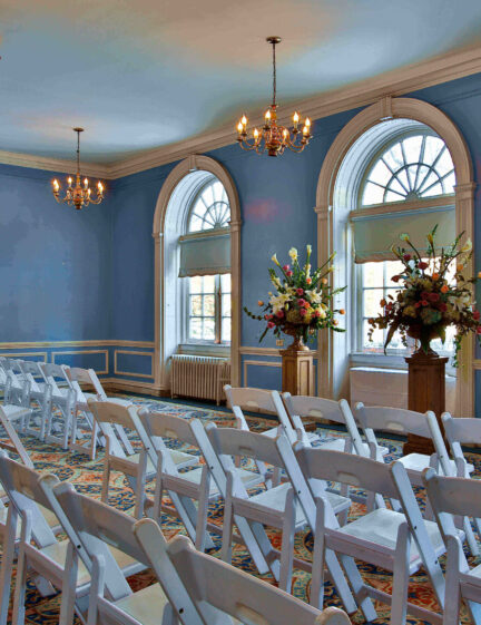 A blue room with chandeliers, arched windows, white chairs in rows, and floral arrangements at the front.