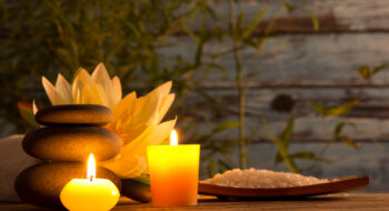 Three stacked stones, candles, a lotus flower, and a tray of bath salts on a wooden surface, creating a spa ambiance.