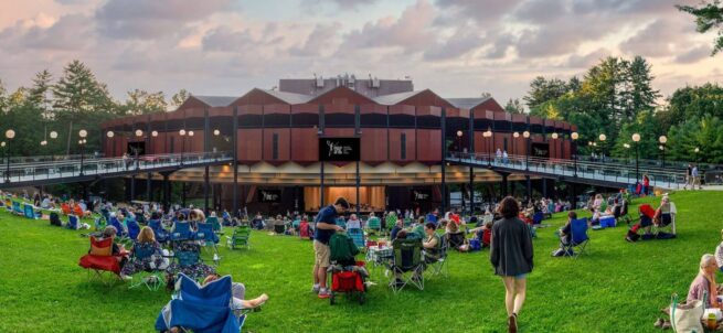People sit on lawn chairs facing an outdoor theater stage at sunset, surrounded by trees and grass.