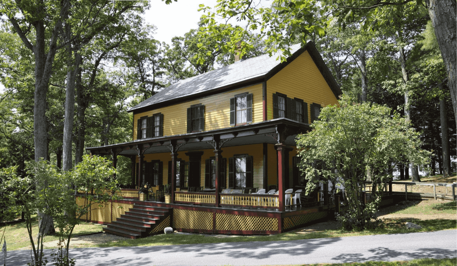 Yellow two-story house with a wraparound porch, surrounded by trees on a sunny day.