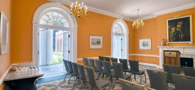 Small meeting room with rows of chairs, chandeliers, orange walls, and open doors leading outside.