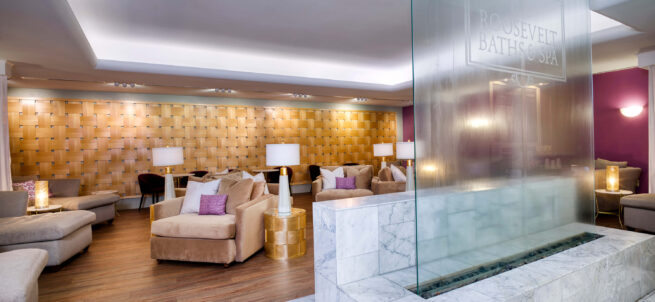 Modern spa lounge with beige sofas, gold accents, and a marble water feature labeled Roosevelt Baths & Spa.
