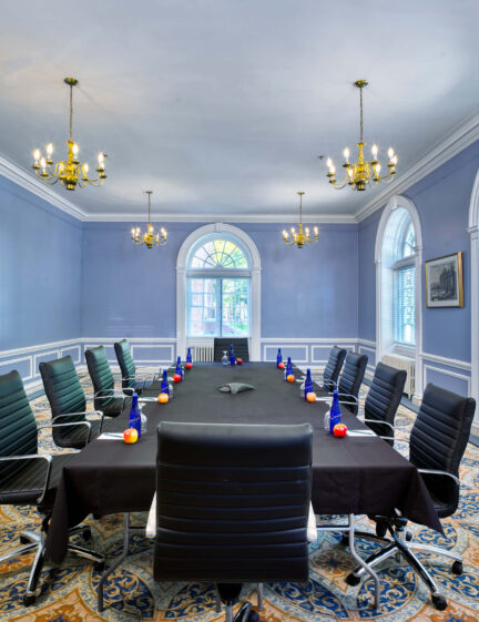 Elegant conference room with a long table, black chairs, chandeliers, and large arched windows.