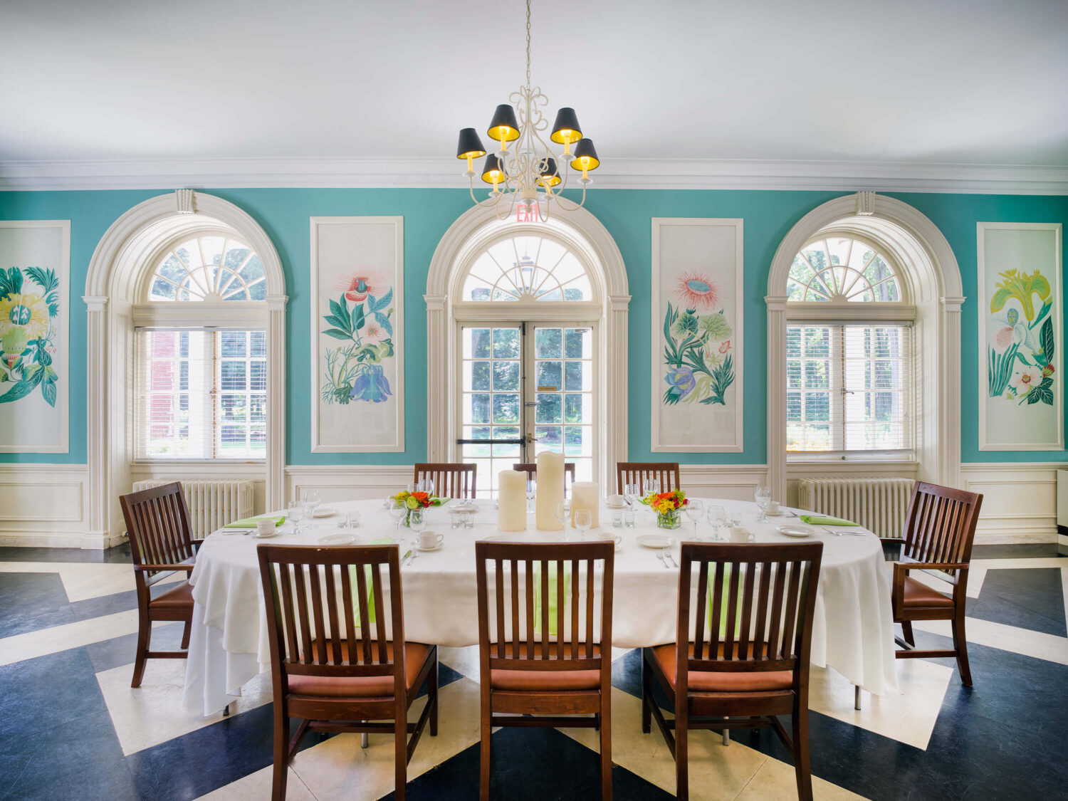 Elegant dining room with a round table, floral wall art, arched windows, and teal walls.