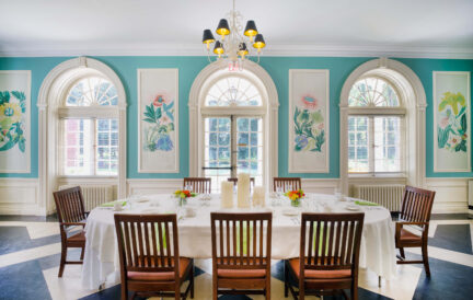 Elegant dining room with a round table, floral wall art, arched windows, and teal walls.