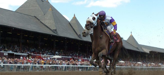 Two jockeys race their horses on a dirt track, with a grandstand full of spectators in the background.