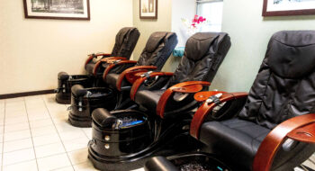 Four black pedicure spa chairs with wooden armrests lined up in a clean, modern salon.
